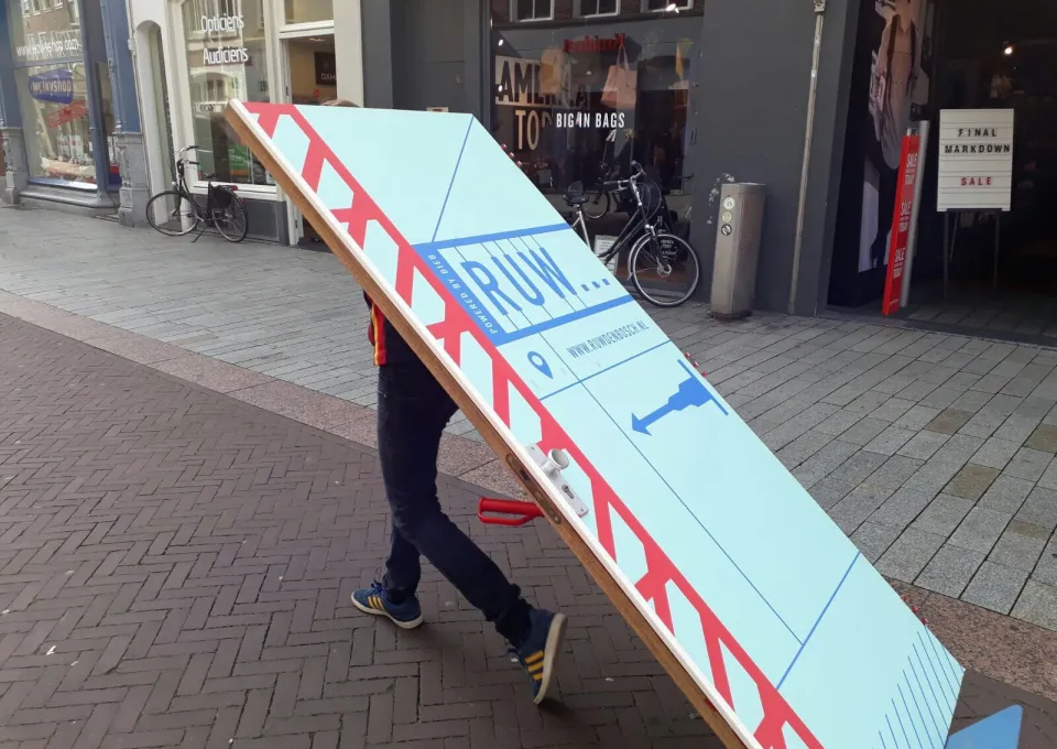 Persoon rijdt met een deur op een steekkarretje door een winkelstraat, met daarop het logo van RUW