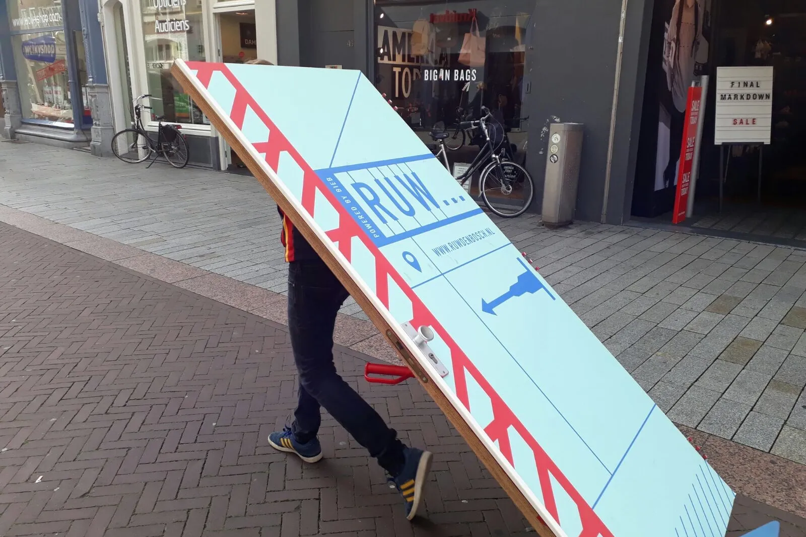 Persoon rijdt met een deur op een steekkarretje door een winkelstraat, met daarop het logo van RUW