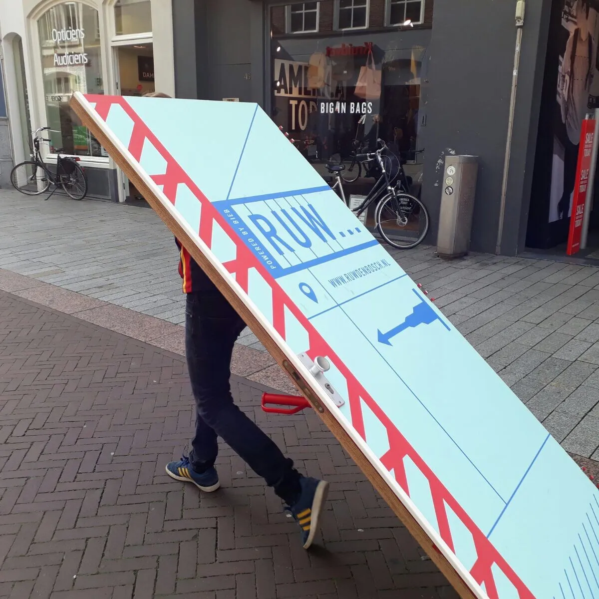 Persoon rijdt met een deur op een steekkarretje door een winkelstraat, met daarop het logo van RUW